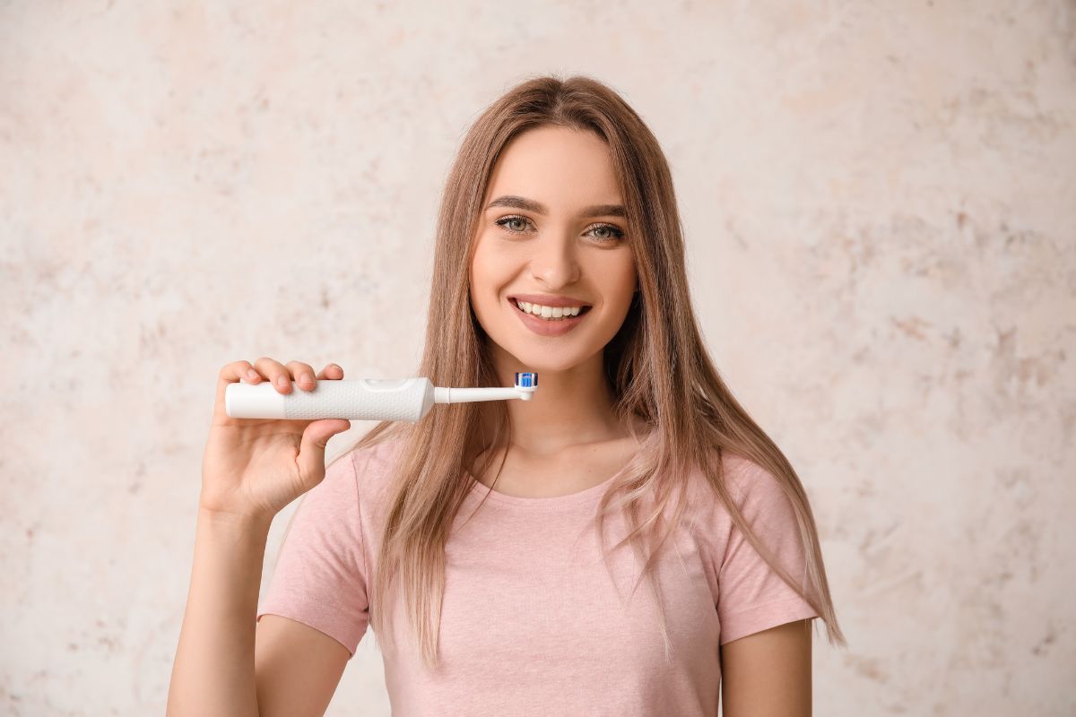 3 Reasons to Pack Your Electric Toothbrush Smiling woman holding toothbrush wearing pink shirt against neutral background